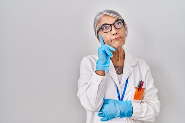 Sticker - Middle age woman with grey hair wearing scientist robe with hand on chin thinking about question, pensive expression. smiling with thoughtful face. doubt concept.