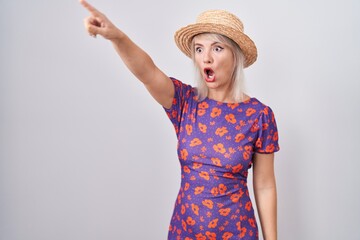 Wall Mural - Young caucasian woman wearing flowers dress and summer hat pointing with finger surprised ahead, open mouth amazed expression, something on the front