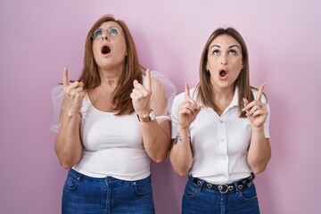 Canvas Print - Hispanic mother and daughter wearing casual white t shirt over pink background amazed and surprised looking up and pointing with fingers and raised arms.