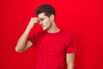 Wall Mural - Young hispanic man standing over red background tired rubbing nose and eyes feeling fatigue and headache. stress and frustration concept.