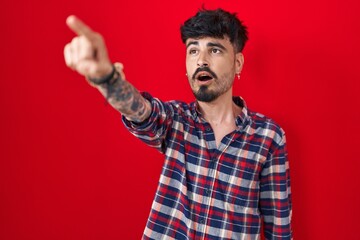 Wall Mural - Young hispanic man with beard standing over red background pointing with finger surprised ahead, open mouth amazed expression, something on the front