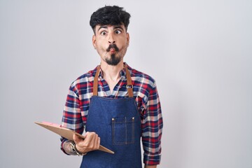 Sticker - Young hispanic man with beard wearing waiter apron holding clipboard making fish face with lips, crazy and comical gesture. funny expression.