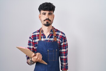 Sticker - Young hispanic man with beard wearing waiter apron holding clipboard relaxed with serious expression on face. simple and natural looking at the camera.