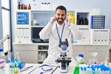 Poster - Young hispanic man with beard working at scientist laboratory covering ears with fingers with annoyed expression for the noise of loud music. deaf concept.