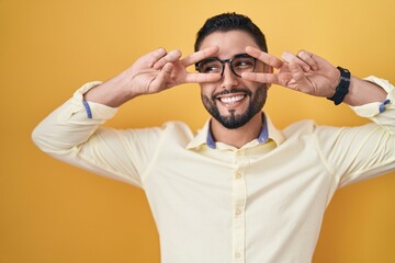 Canvas Print - Hispanic young man wearing business clothes and glasses doing peace symbol with fingers over face, smiling cheerful showing victory