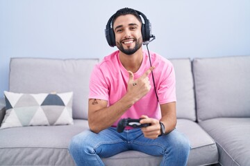 Sticker - Hispanic young man playing video game holding controller sitting on the sofa cheerful with a smile on face pointing with hand and finger up to the side with happy and natural expression