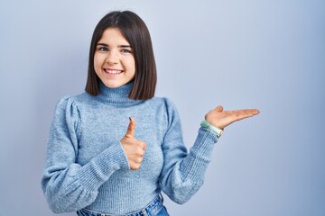 Young hispanic woman standing over blue background showing palm hand and doing ok gesture with thumbs up, smiling happy and cheerful