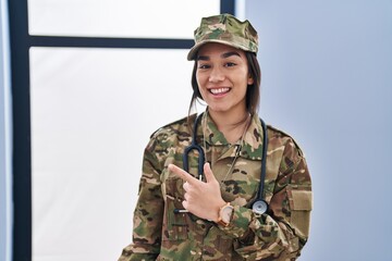 Wall Mural - Young south asian woman wearing camouflage army uniform and stethoscope smiling cheerful pointing with hand and finger up to the side