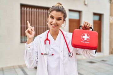 Poster - Young doctor woman holding first aid kit smiling happy pointing with hand and finger to the side