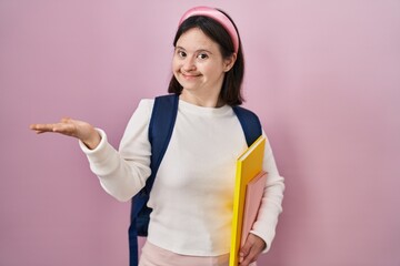 Poster - Woman with down syndrome wearing student backpack and holding books smiling cheerful presenting and pointing with palm of hand looking at the camera.