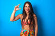 beautiful brunette woman wearing swimwear over blue background smiling and confident gesturing with hand doing small size sign with fingers looking and the camera. Measure concept