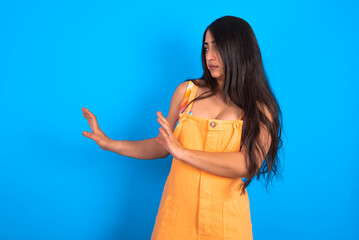 Displeased brunette woman wearing orange overalls over blue studio background keeps hands towards empty space and asks not come closer sees something unpleasant