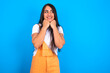 © Jihan - brunette woman wearing orange overalls over blue studio background  holds hands under chin, glad to hear heartwarming words from stranger