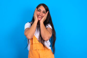 Wall Mural - Happy brunette woman wearing orange overalls over blue studio background touches both cheeks gently, has tender smile, shows white teeth, gazes positively straightly at camera,