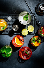 Cocktails set on black bar counter, top view. Mixology concept. Assortment of colorful strong and low alcohol drinks for cocktail party. Dark background, bar tools, hard ligh