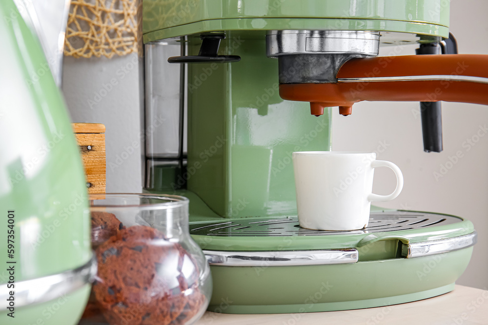 Modern coffee machine, cups and jar with cookies on table near white wall, closeup