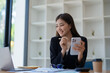 © Jirapong - Portrait of a young Asian woman showing a smiling face as she uses her phone, computer and financial documents on her desk in the early morning hours