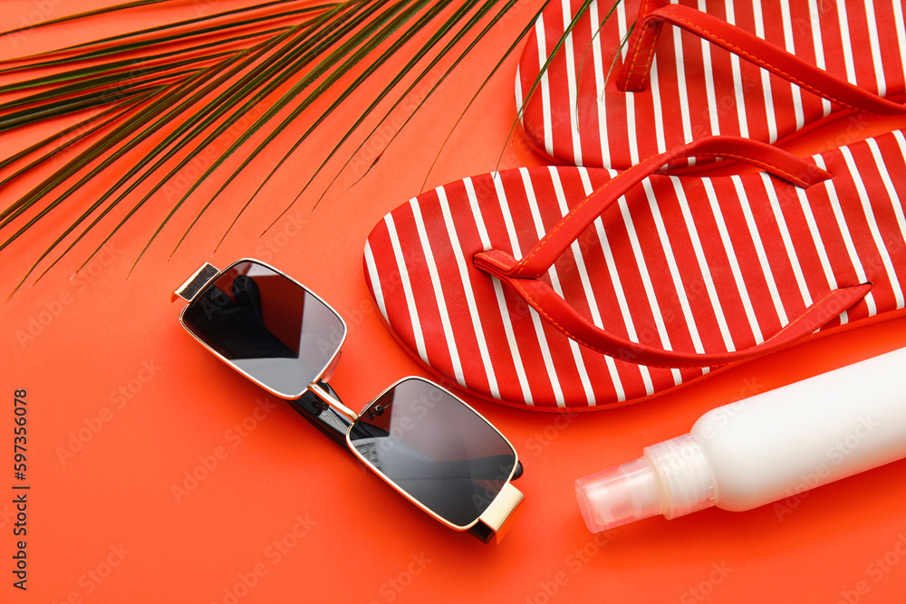 Flip-flops, sunglasses, bottle of sunscreen and palm leaf on red background