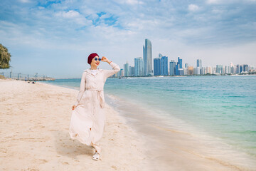 Wall Mural - Young woman walks along sandy beach and takes occasional glances toward the impressive skyline, admiring the architectural design and marveling at the scale of Abu Dhabi city's development, UAE