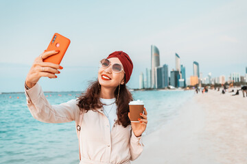 Wall Mural - With the crystal clear waters and towering skyscrapers of Abu Dhabi in the background, a young girl takes a selfie on her smartphone on the beach, capturing the moment and the stunning view.