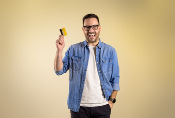 Handsome male customer with hand in pocket showing credit card and laughing ecstatically on isolated beige background. Cheerful young man recommending online shopping and cashless payment