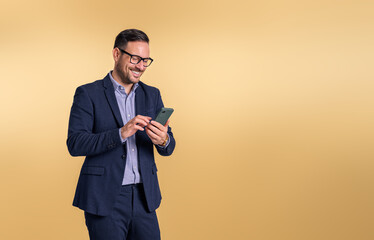 Young handsome businessman dressed in elegant suit messaging over mobile phone. Male professional manager smiling and checking social media while standing isolated on beige background