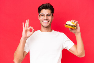 Wall Mural - Young Caucasian man holding a burger isolated on red background showing ok sign with fingers