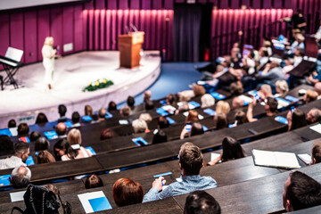 Wall Mural - Business and entrepreneurship symposium. Female speaker giving a talk at business meeting. Audience in conference hall. Rear view of unrecognized participant in audience.