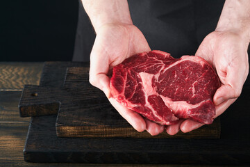 Wall Mural - Chef cutting steak beef ribeye. Mans hands hold raw steak Ribeye on rustic wooden cutting board on black background. Cooking, recipes and eating concept. Selective focus.