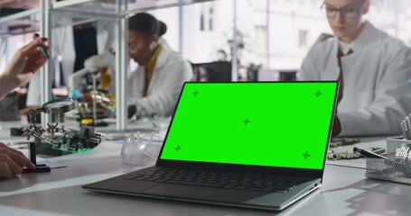 Fototapete - Laptop Computer with Green Screen Mock Up Display Standing on a Table in a Science Class Laboratory. Diverse Students Practicing Soldering Electronic Components for Their Tech Projects