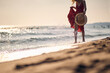 © luckybusiness - Young woman in waving red summer dress walking on beach holding fashionable straw hat. Vacation, holiday, lifestyle concept.