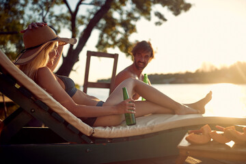 A young couple in swimsuit sitting on sunbeds and enjoying the sun and drink on the river bank. Summer, river, vacation