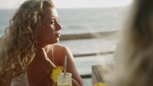 Blonde Young Woman In White Dress In A Sea View Tropical Cafe Talks, Smiles, Wind Blows Her Long Hair. Happy Young Curly Female Chats With Girls In Seaside Outdoor Restaurant At Sunset. Ocean View Bar