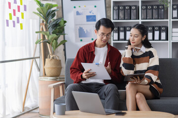 Young asian successful woman and man discussing paperwork and using a laptop together sitting on a sofa in a modern office.