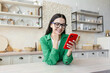 © Liubomir - Young woman standing in kitchen at home, leaning on table and using mobile phone, chatting, checking social networks, sending messages, texting with friends, boyfriend.