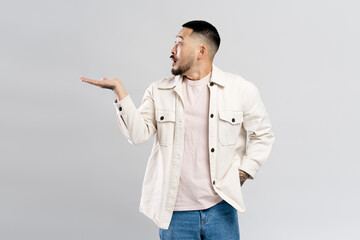 Wall Mural - Surprised attractive asian man pointing with hand at copy space, isolated on gray background