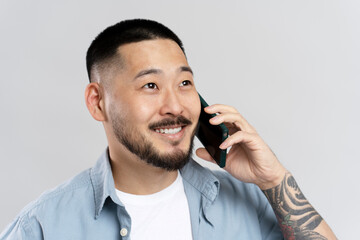 Wall Mural - Portrait of handsome Asian man with stylish tattoo talking on mobile phone, isolated on gray background. Successful businessman answering call looking away 