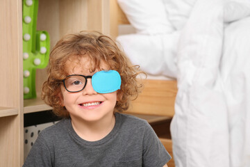 Happy boy with eye patch on glasses near shelf indoors. Strabismus treatment