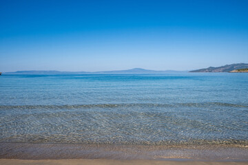 Wall Mural - Sandy empty beach transparent wavy sea water clear blue sky background. Greek seascape, sunny day.