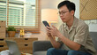 © Prathankarnpap - Image of adult asian man in casual watching video or playing game on smart phone, sitting on couch at home