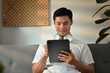 © Prathankarnpap - Handsome Asian man sitting on couch working remotely online on digital tablet at home