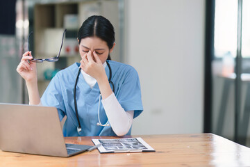 Professional millennial female doctor or nurse working on laptop computer and clipboard with a tense expression while sitting at a desk in a hospital consultation room.