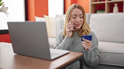 Poster - Young blonde woman talking on smartphone holding credit card sitting on sofa at home