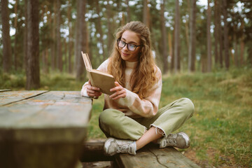 Happy young woman is sitting at table with book in beautiful forest. Beautiful woman is enjoying fresh air in forest reading book. Concept of relaxation, solitude with nature, lifestyle.