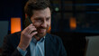 © Yuliia - Caucasian man sit at desk evening night office work online on computer answer mobile call adult middle-aged businessman 40s male smiling entrepreneur talking phone distant business talk by smartphone