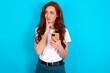 © Jihan - Thinking dreaming young redhead woman wearing white T-shirt over blue background using mobile phone and holding hand on face. Taking decisions and social media concept.