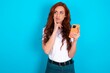 © Jihan - Image of a thinking dreaming young redhead woman wearing white T-shirt over blue background using mobile phone and holding hand on face. Taking decisions and social media concept.