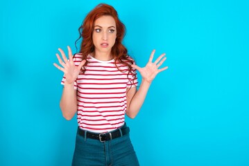 Wall Mural - young redhead woman wearing striped T-shirt over blue background shouts loud, keeps eyes opened and hands tense.
