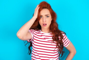 Wall Mural - Embarrassed young redhead woman wearing striped T-shirt over blue background with shocked expression, expresses great amazement, Puzzled model poses indoor