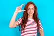 © Roquillo - young redhead woman wearing striped T-shirt over blue background smiling and confident gesturing with hand doing small size sign with fingers looking and the camera. Measure concept